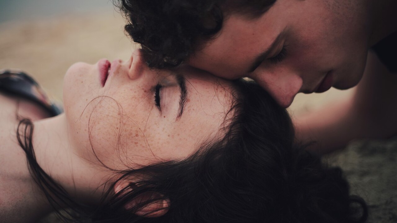 Two men, both with their eyes closed, are kissing the woman on the forehead, and she is lying on the ground