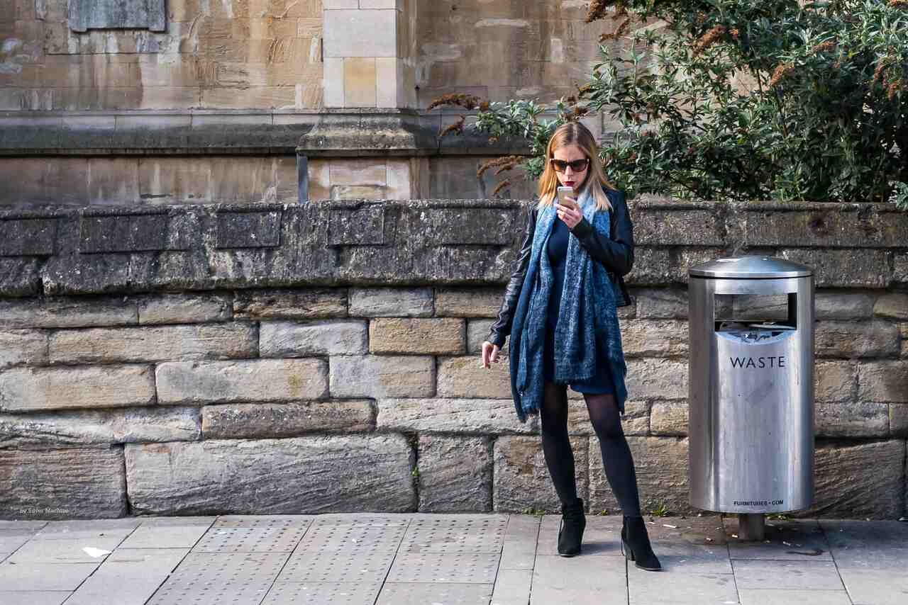 A woman is standing on the street, holding her phone and smoking a cigarette. She is reading her messages.